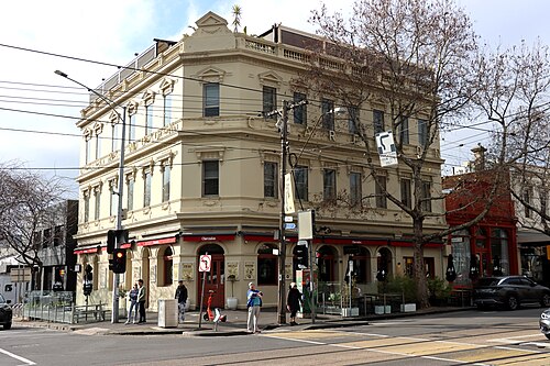 A three storey rendered Victorian corner hotel with CLARENDON in relief lettering across the parapet, arched ground floor windows, a red awning over the footpath, and pedestrians crossing at the corner.