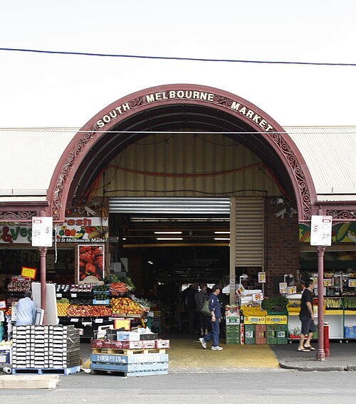 The wrought iron arched entrance canopy of the market, lettered SOUTH MELBOURNE MARKET, with fruit and vegetable stalls and shoppers passing beneath.