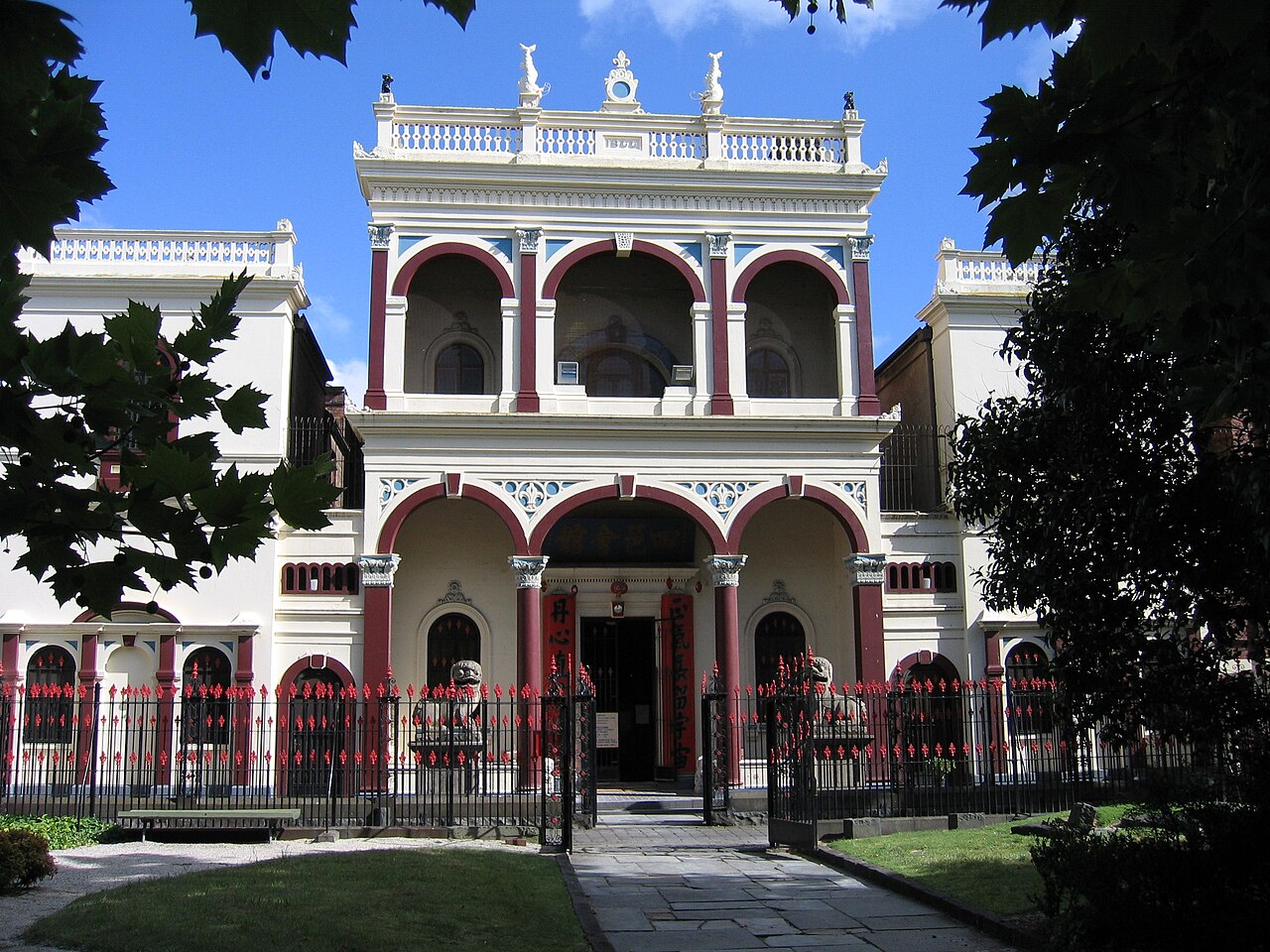 The cream and red rendered facade of the temple, twin arched loggias with a balustraded parapet, stone lion statues behind a red spear topped iron fence.