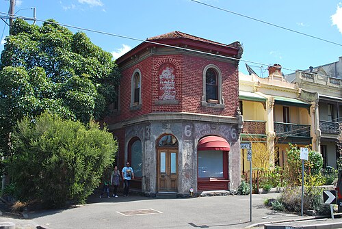 A two storey red brick corner shop with a splayed corner bay, arched window heads, faded painted advertising lettering on the brickwork, and a red awning over the recessed door, a Bank St street sign at the kerb.