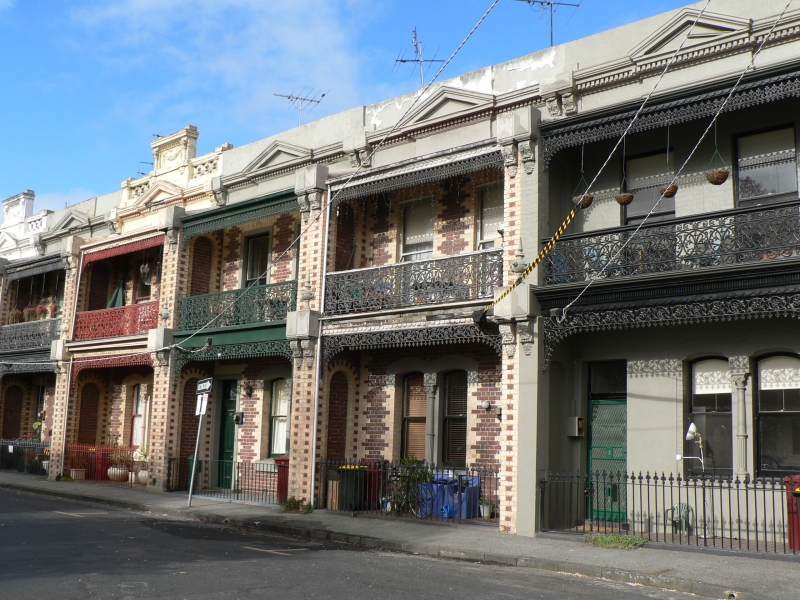 A row of two storey polychrome brick Victorian terrace houses with ornamental cast iron lace verandah balconies and parapets along a quiet residential street.