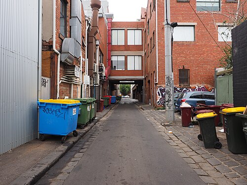 A narrow laneway paved in bluestone pitchers between red brick warehouse buildings, wheelie bins along both kerbs, graffiti tags on the brick, and a loading bridge crossing overhead.