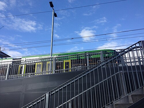 A green tram behind metal security railings and stairs leading up to a raised platform, against a clear sky.