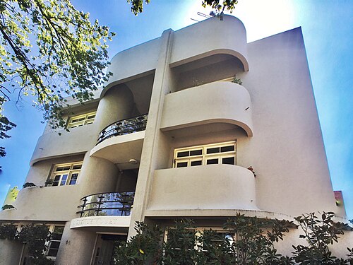 A three storey cream rendered Art Deco apartment building with rounded balconies and metal balustrades, seen from street level.