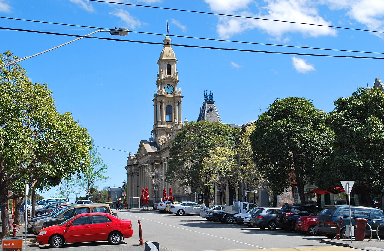 Bank Street with a parked car in the foreground, trees framing a view down the street to the South Melbourne Town Hall's clock tower and slate turret in the distance, a Bank St street sign at the kerb.