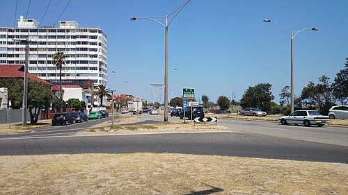 Marine Parade running along the coast, a white mid rise apartment building at the intersection under a clear sky.