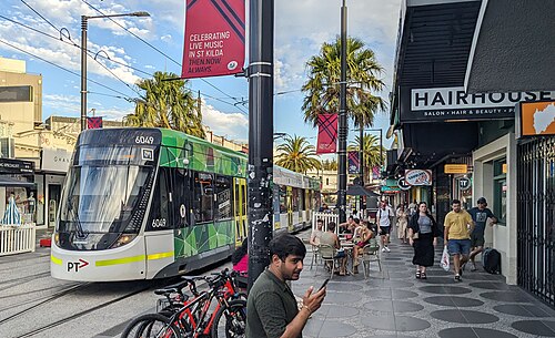 A green E class tram pulling away from the Acland Street terminus, shopfronts and outdoor tables along the kerb.