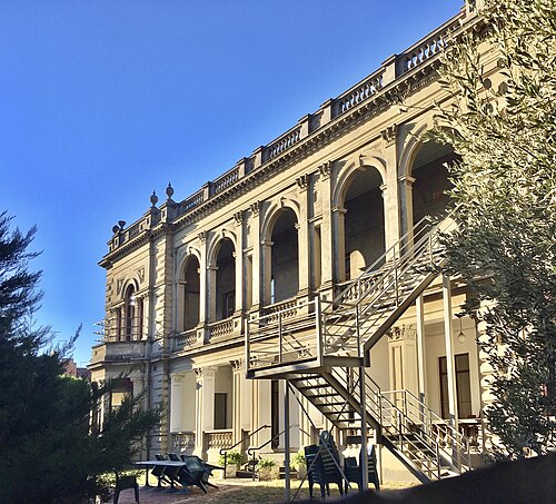 Eildon Mansion's rear elevation, an arched loggia and balustrades on a two storey rendered nineteenth century mansion, with a metal fire escape stair added later.