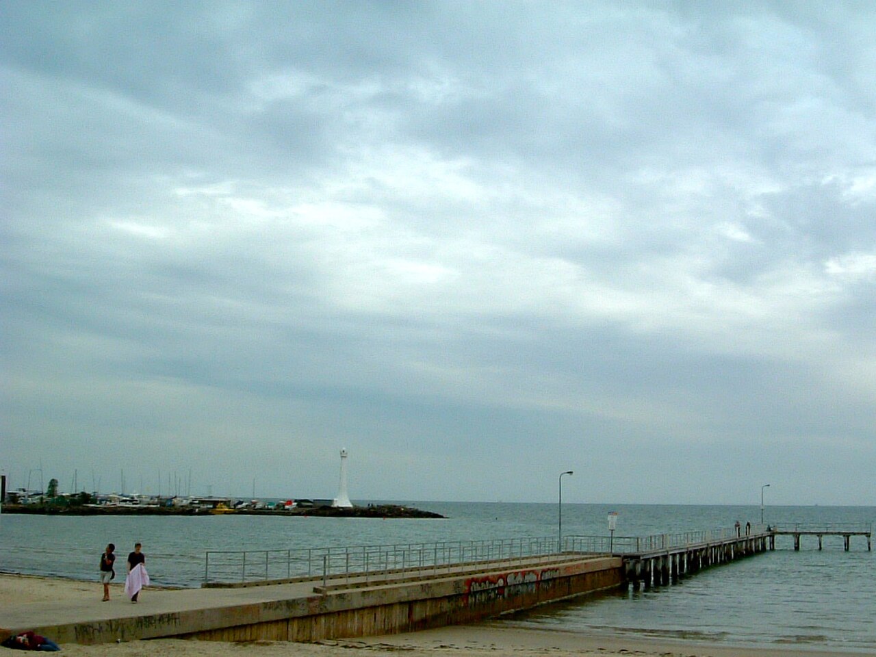 A long wooden pier running out into Port Phillip Bay under an overcast sky, a white light tower at its far end.