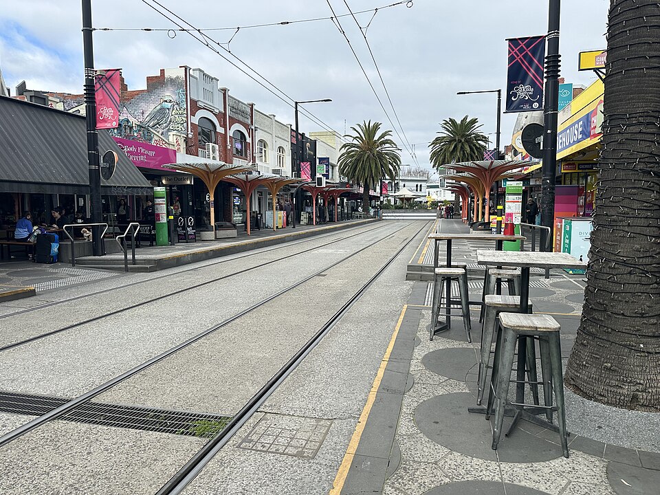 Acland Street's pedestrianised cake shop strip, striped awnings and outdoor cafe tables either side of the tram tracks.