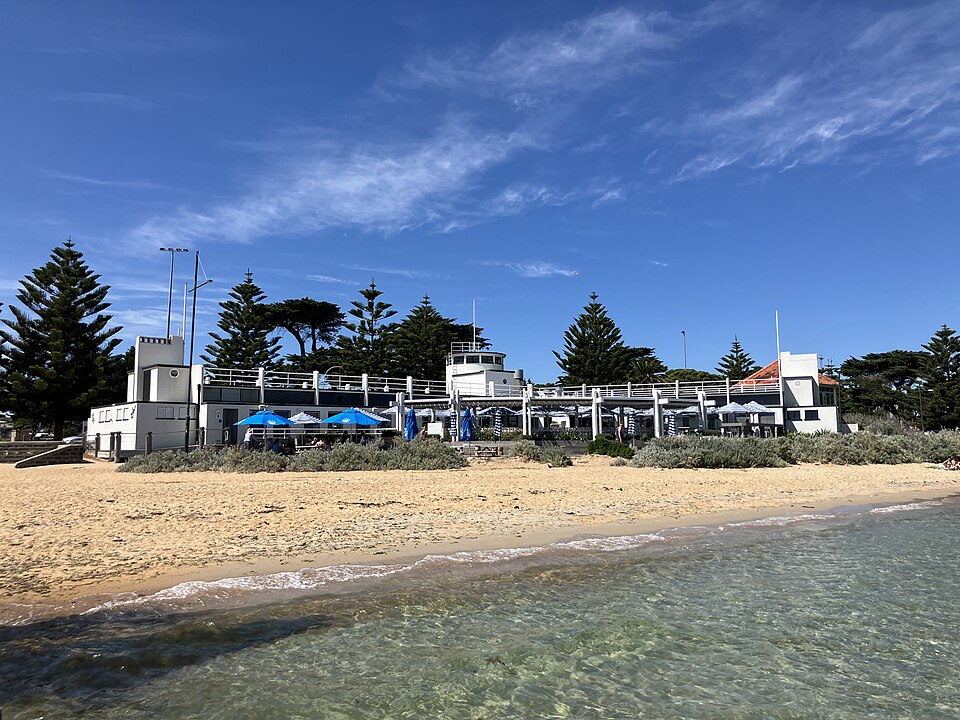 The Williamstown Beach Dressing Pavilion, a white painted Art Deco building with a central tower and open terrace on the Esplanade, at the other end of Williamstown Beach from the Life Saving Club building the scene names.