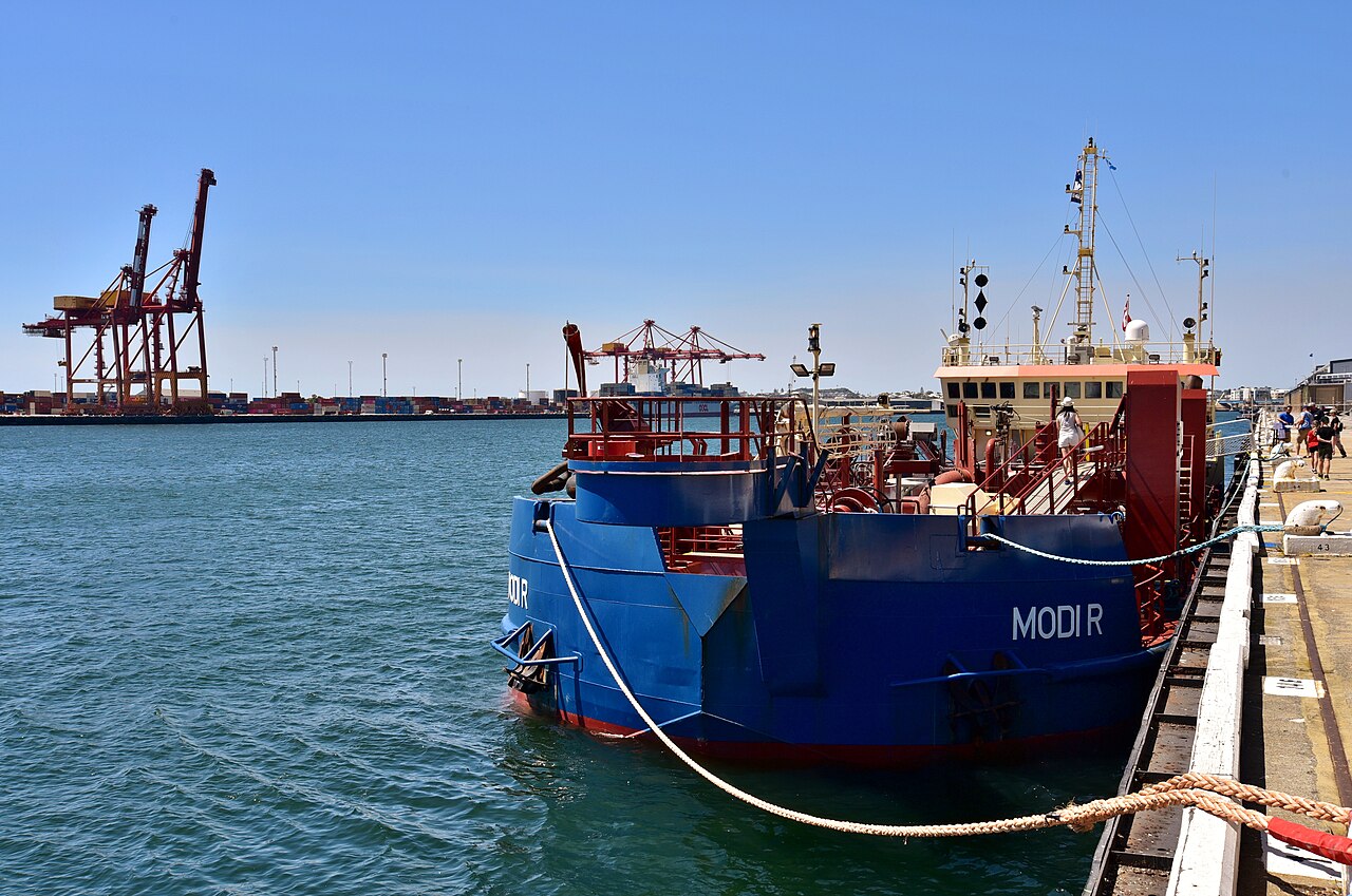 The dredge Modi R tied up at the quay, with the container terminal's red gantry cranes and stacked boxes on the far side of the water.