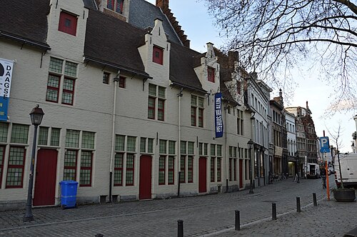 The Huis van Alijn on the Kraanlei, a whitewashed almshouse with red doors and stepped dormers, with a blue banner hung on the front and bollards along the cobbles.