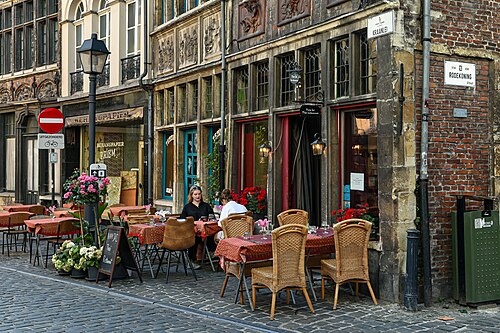 The Kraanlei at the corner of Rodekoningstraat, with restaurant tables out on the cobbles and BEHANGPAPIER PRIEM painted across a shop window. The carved stone front on the left is the Seven Works of Mercy house.