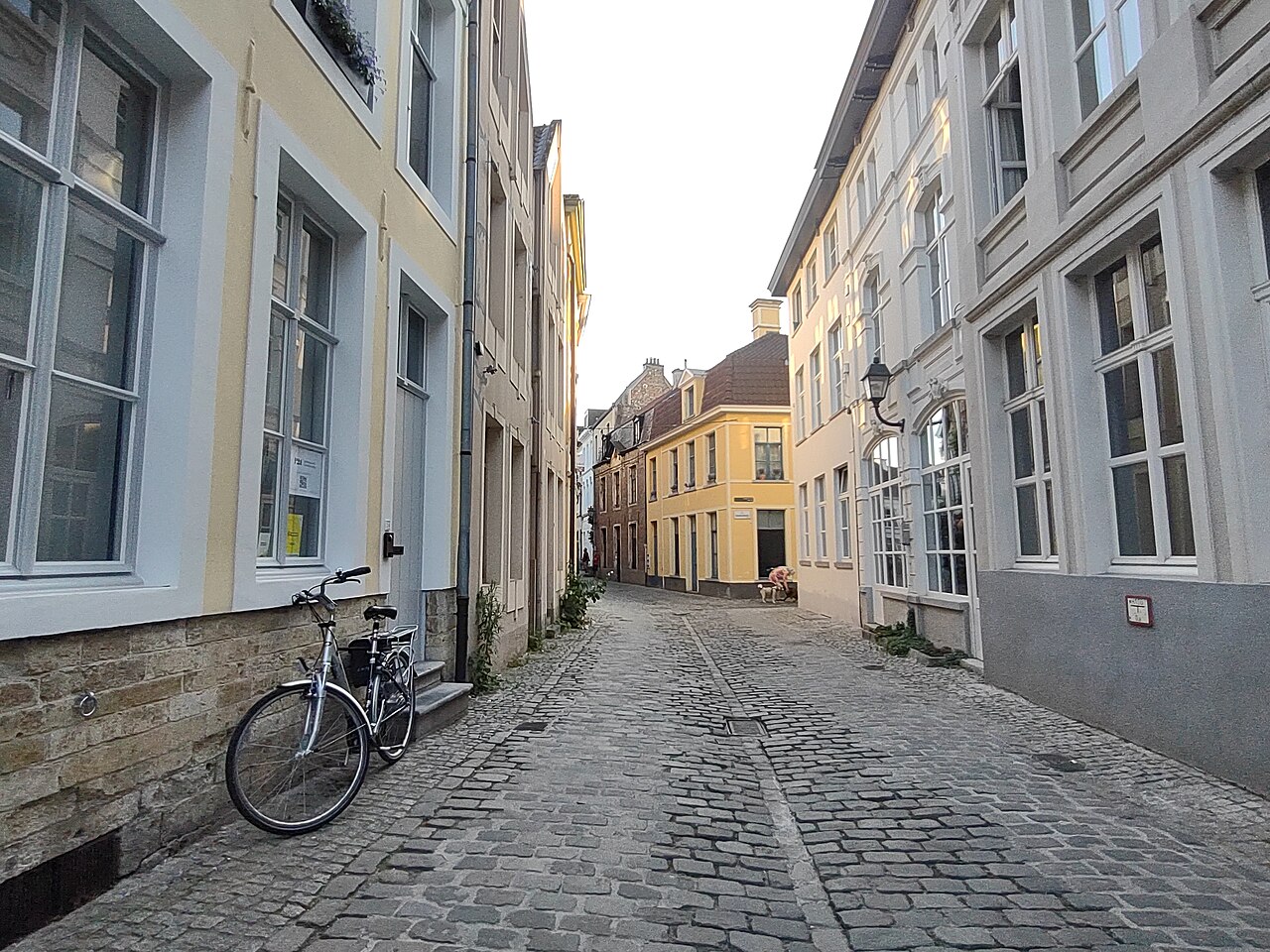The Plotersgracht under a white sky, a cobbled lane between three storey rendered houses, with a bicycle against the wall on the left and a yellow house at the far end.