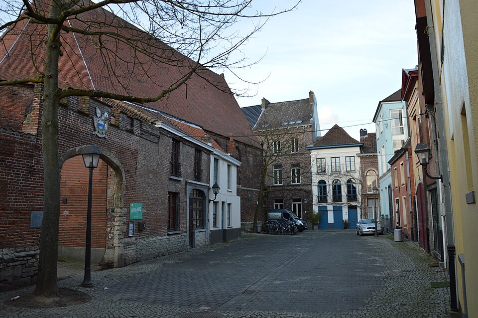 The Kaatsspelplein, an open paved square with the long tiled roof of the Caermersklooster along the left and a red brick arch with a painted coat of arms above it.
