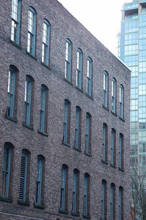 The upper storeys of a Mainland Street warehouse, seen looking up from the sidewalk, round headed windows in blue trim rising against a newer glass tower just behind.