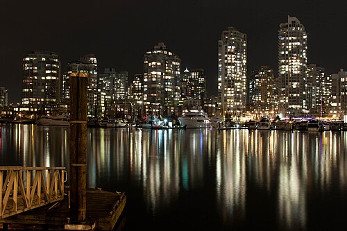 Yaletown's waterfront towers lit up after dark, reflected in False Creek, with yachts and a houseboat moored along the shore.