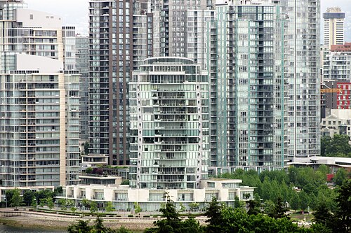 A curved glass residential tower rising above a row of townhouses on the Yaletown side of False Creek, seen from across the water.