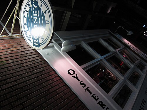 Rodney's Oyster House's round sign mounted over the entrance on Hamilton Street, seen from below at night, the brick wall lit by an awning lamp.