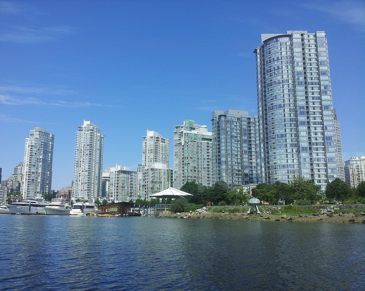 A row of glass residential towers along the Yaletown side of False Creek, seen from the water, with a marina of yachts and houseboats in the foreground.