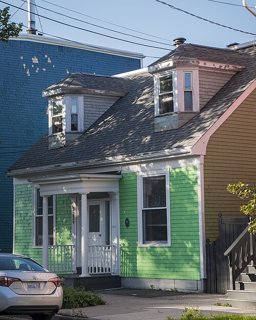 The cottage at 2229 Brunswick Street, bright green shingles under a shingled roof with two dormer bay windows and a small white porch, a mustard yellow house against it on the right and a blue painted wall behind.