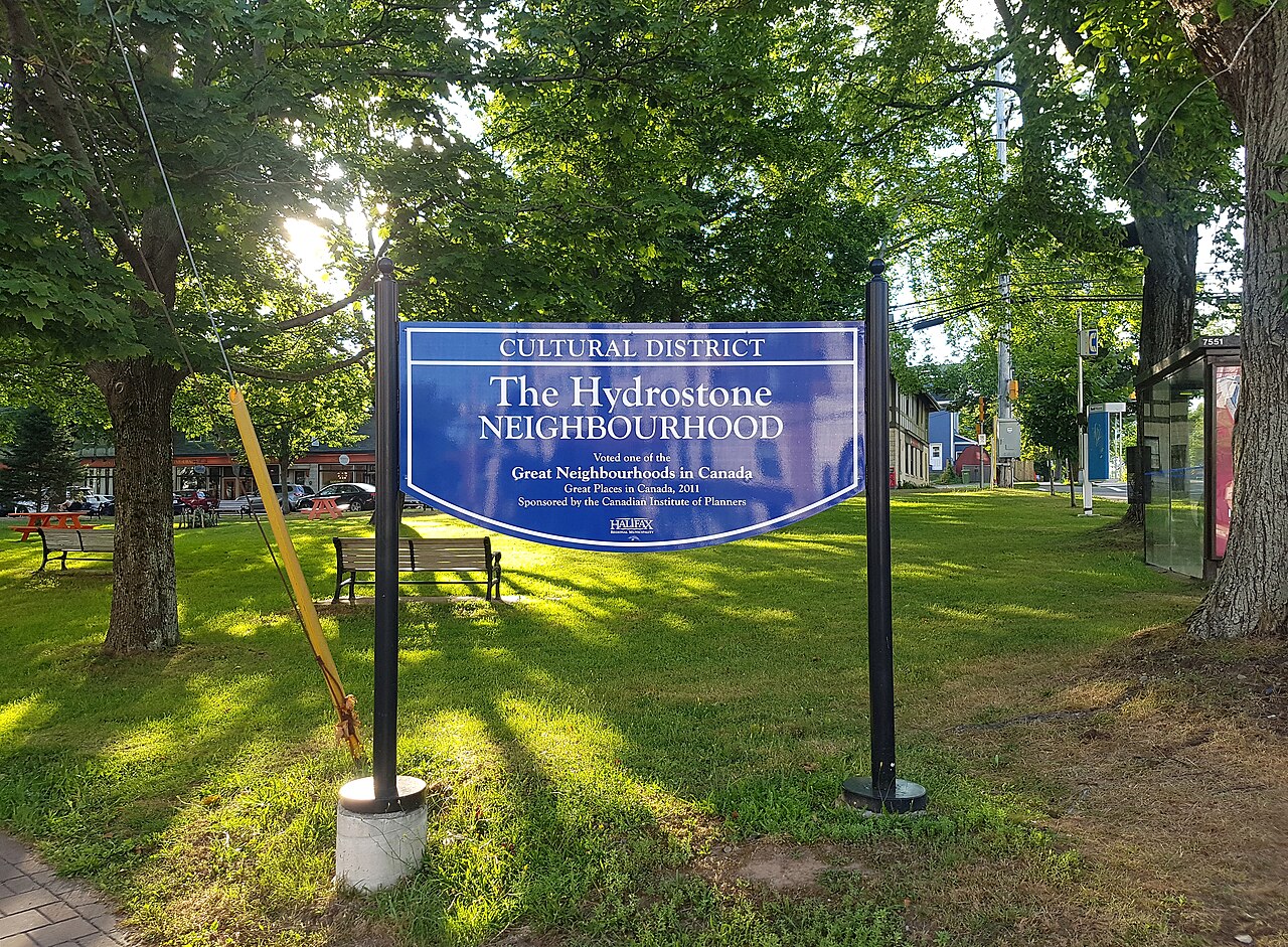 The Hydrostone Neighbourhood sign in the grass on Young Street, with picnic tables and a bench under the trees and the market storefronts across the road behind them.