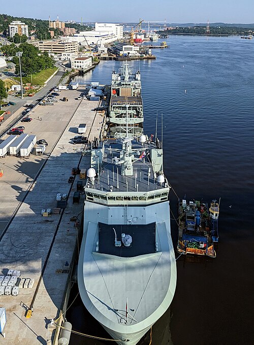 Two grey naval ships alongside the jetty at HMC Dockyard, seen from above and looking north up the Narrows, with the Halifax Shipyard assembly hall on the shore and the MacKay Bridge beyond it.
