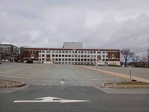 Building S-15 at CFB Halifax, a long four storey block in brick and white render seen across an empty parking lot under grey cloud.