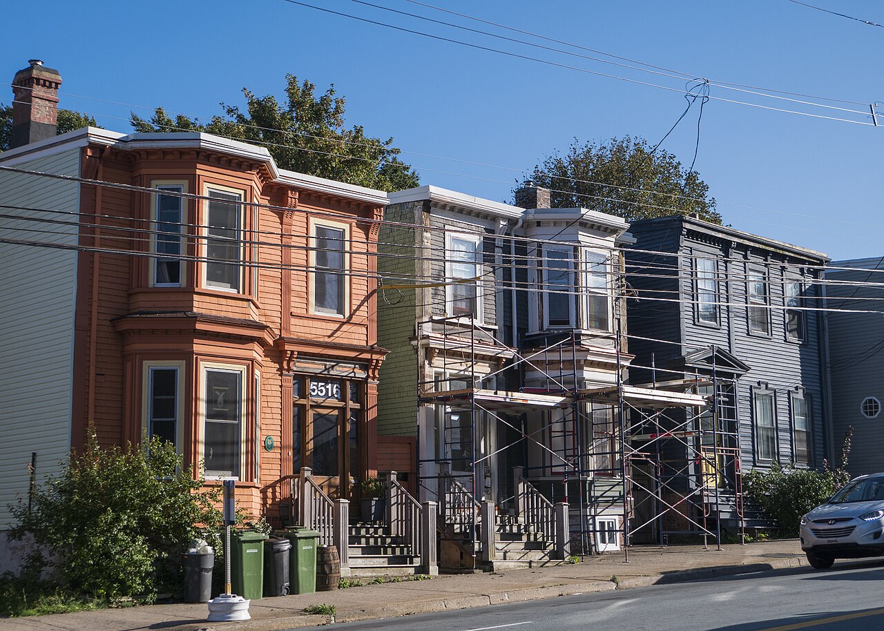 A run of two storey wooden houses on North Street, faced in shingle and clapboard and painted orange, olive and grey, with tiered bay windows and front steps that reach the sidewalk. One house is behind scaffolding.