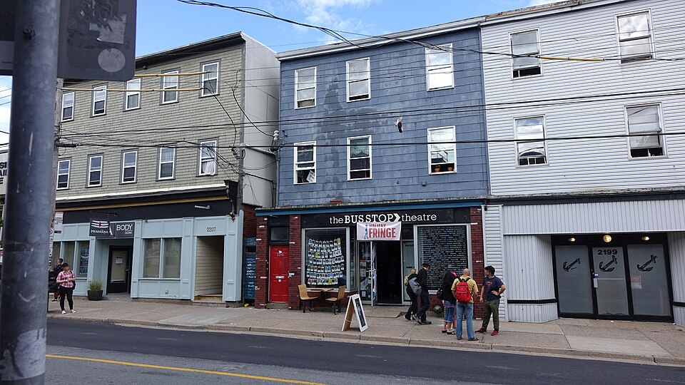 Gottingen Street outside the Bus Stop Theatre at 2203, three storey wood clad buildings with storefronts at street level, roll shutters painted with anchors at 2199 and people talking on the sidewalk.