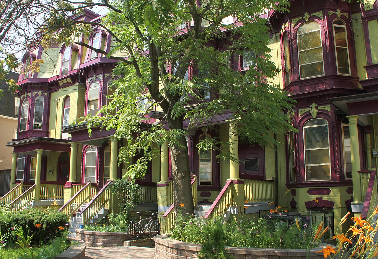 A row of Victorian houses painted olive and maroon, with wraparound porches, decorative brackets and bay windows, a mature tree and orange daylilies in front.