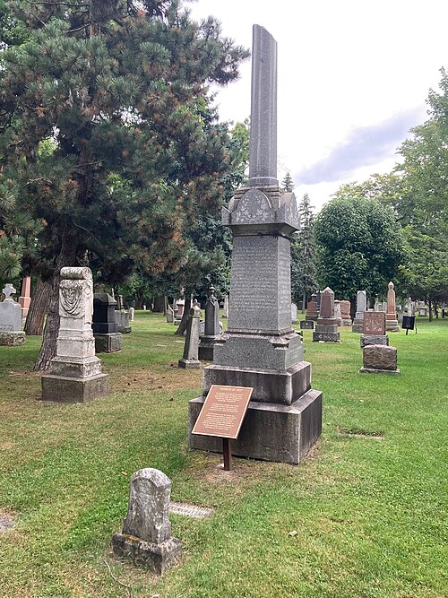 The Patriots of 1837 monument at the Toronto Necropolis, a granite obelisk on a stepped base among older headstones and pine trees.