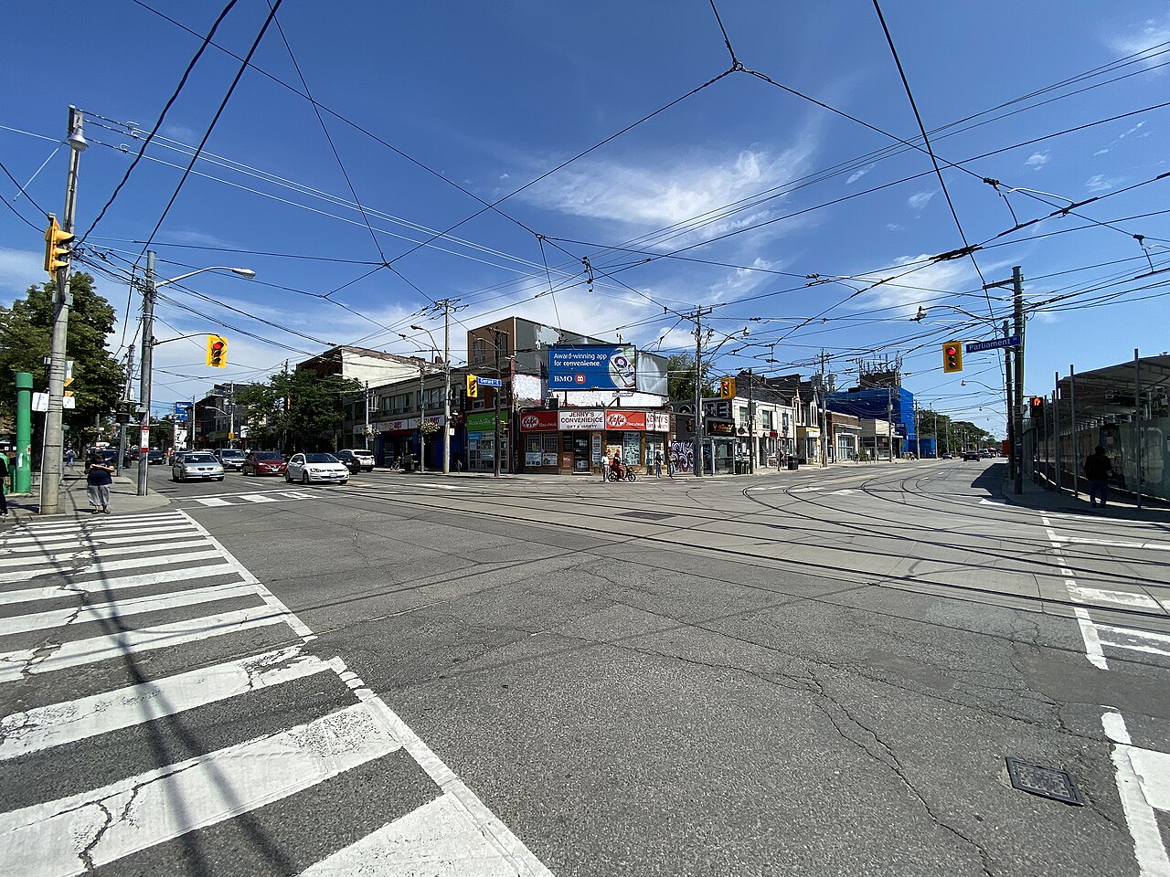 Gerrard Street East at Parliament Street, a wide sunlit intersection with streetcar tracks, a corner convenience store and low brick storefronts, a Parliament street sign overhead.