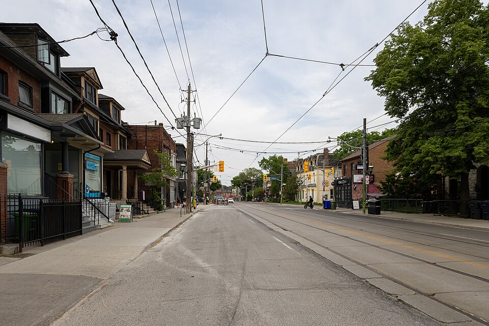 Carlton Street looking east near Ontario Street, brick rowhouses and storefronts on both sides, streetcar wires overhead and a cyclist riding down the middle of the road.