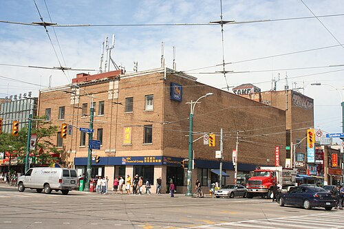 The three storey brick building on the northeast corner of Spadina Avenue and Dundas Street West, now a bank branch, seen from across the intersection with a fire truck and traffic passing.