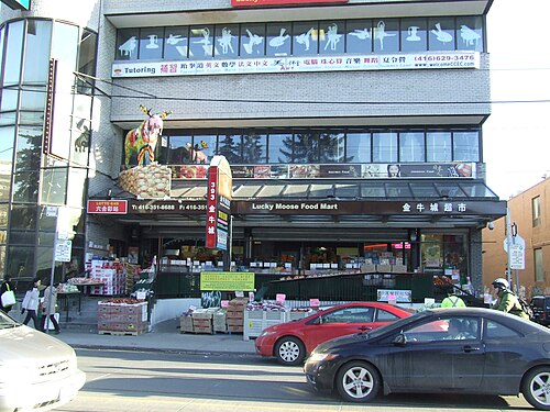 Lucky Moose Food Mart, a grocery store with crates of produce stacked along the sidewalk under its awning and cars parked at the curb.