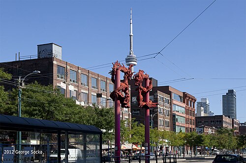 Two red dragon figures wound around lamp posts at a streetcar stop on Spadina Avenue, with the CN Tower visible above the rooftops behind them.