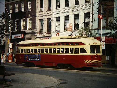 A red and cream PCC streetcar stopped near Spadina Avenue and Dundas Street West in 1983, brick storefronts and a Scotiabank branch behind it.