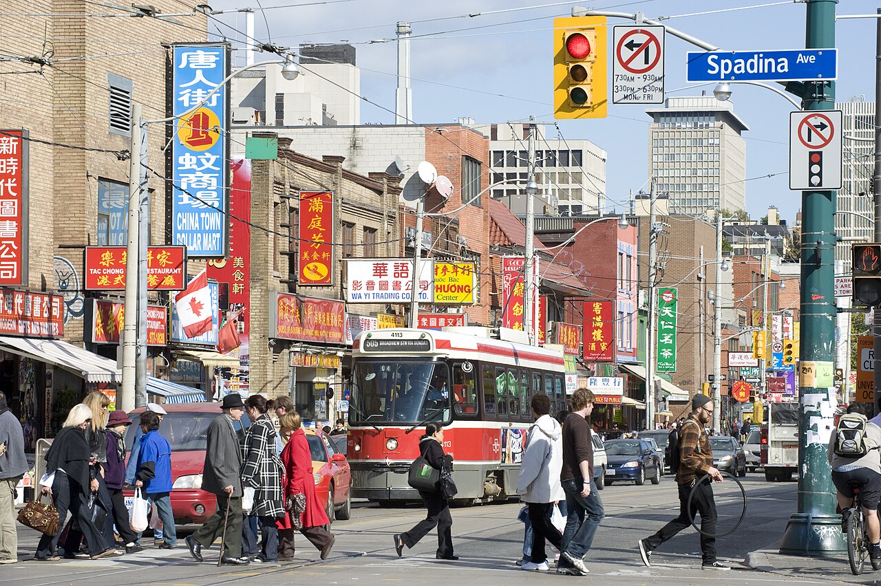 Dundas Street West at Spadina Avenue, a red streetcar crossing the intersection with pedestrians in front of it, a green Spadina Ave street sign, and red vertical shop signs in Chinese and English lining the block.
