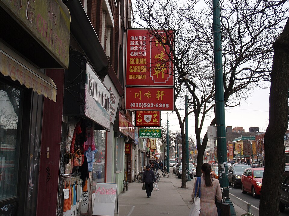 A row of Chinatown storefronts along a sidewalk lined with bare trees, red signboards including one for the Sichuan Garden Restaurant, and pedestrians carrying shopping bags.