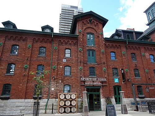 A four story brick warehouse with a stepped gable and an arched window at its center, green painted window frames and doors, and a stack of wooden barrels by the entrance.