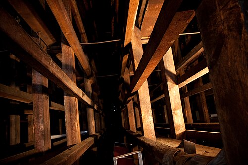 The inside of a barrel aging room, rows of wooden support beams running back into the dark with a single work light glowing partway down the row.