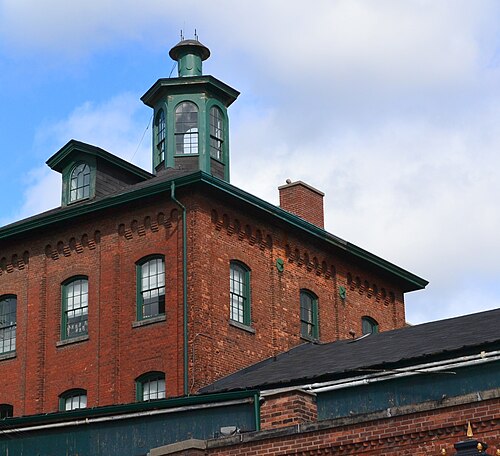 The peaked green roof and cupola of a brick building, corbelled brickwork under the eaves and a brick chimney beside it.