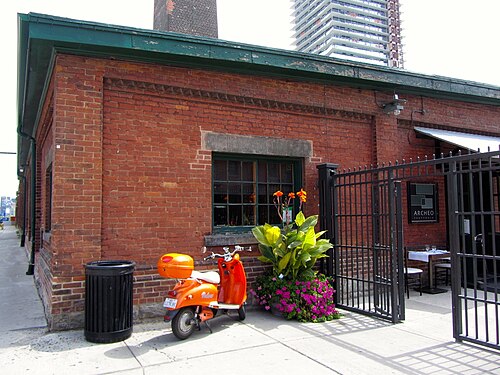 The corner of a low brick building with a stepped parapet, a green iron gate open onto a restaurant patio, and a parked scooter beside a trash can and a planter of flowers.