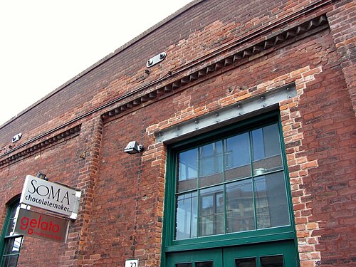 A brick wall with corbelled brick detailing under the roofline and a green painted door and window, a chocolate shop's sign mounted on the wall beside it.
