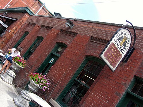 A brick building with arched, green framed windows and a hanging sign over the sidewalk, stone planters of flowers along the front.