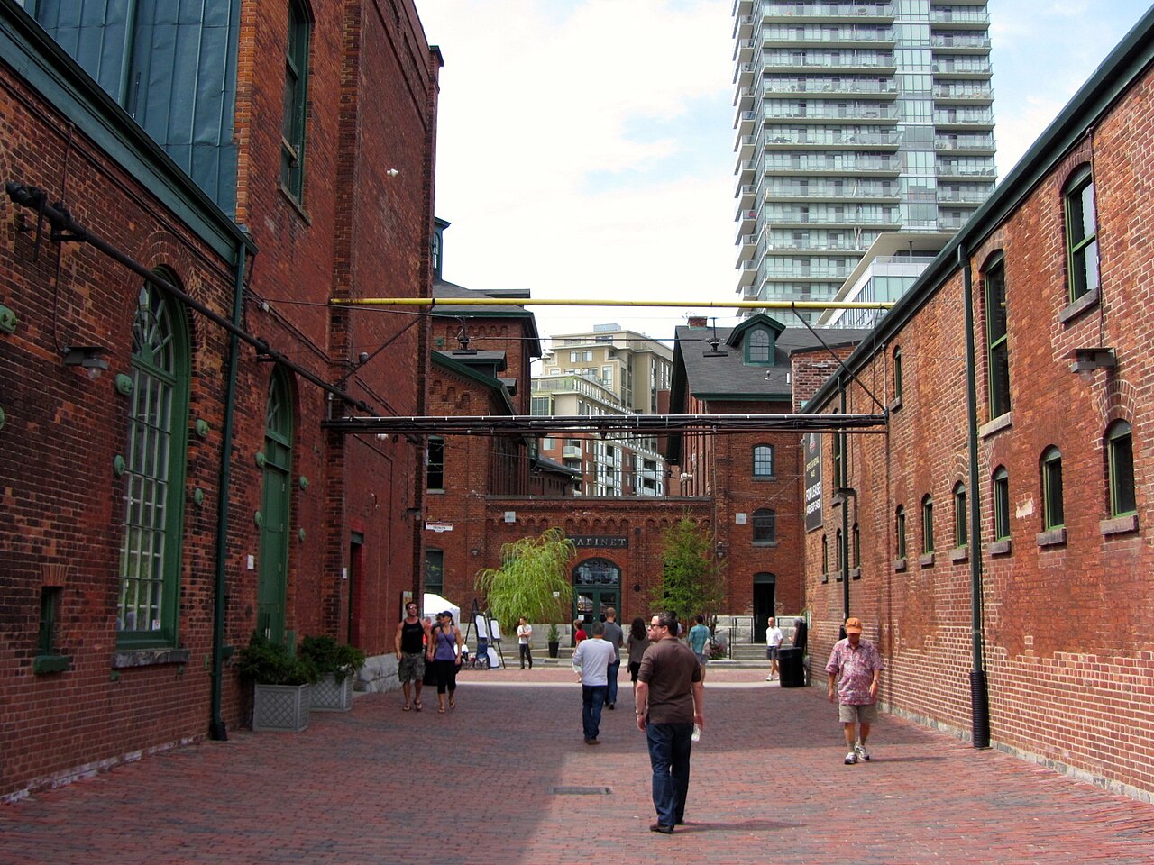 A cobblestone lane between two brick warehouse buildings, with a green iron gas lamp and potted plants, pedestrians walking down toward a cross street lined with more brick buildings and a glass condo tower rising behind them.