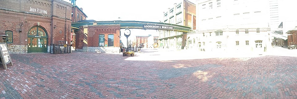 An empty cobblestone lane between brick buildings, a Gooderham and Worts sign mounted on an overhead footbridge and a clock partway down the block.