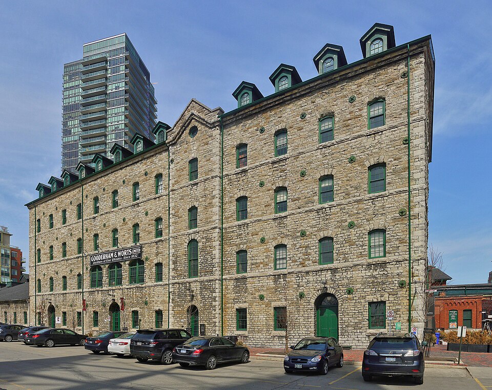 The former Stone Distillery and grist mill, a long stone building with green dormer windows along its roofline and a Gooderham and Worts company sign on the wall, seen across a parking area.